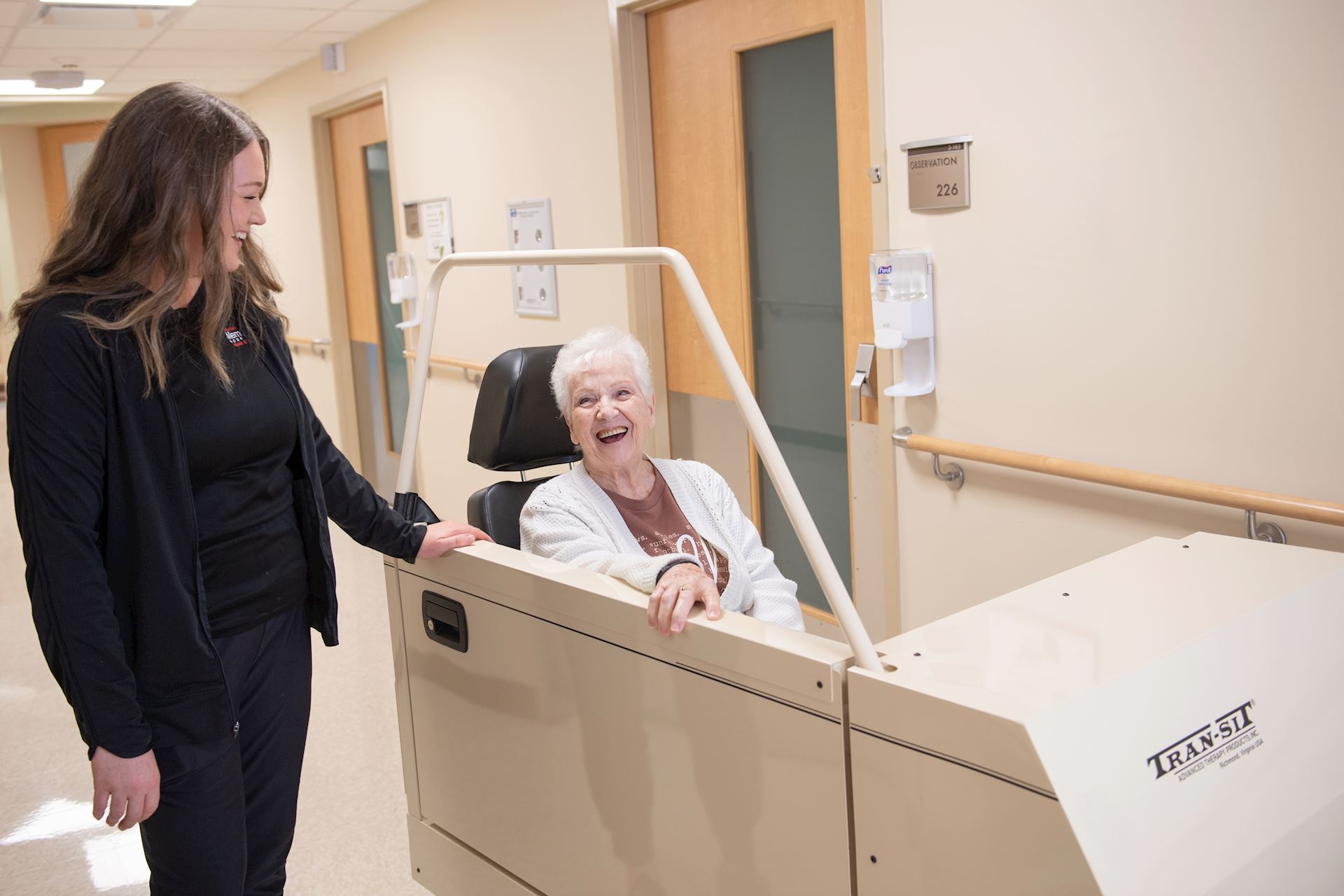 Photograph of a healthcare worker assisting an elderly woman seated in a beige transit wheelchair in a hospital hallway.