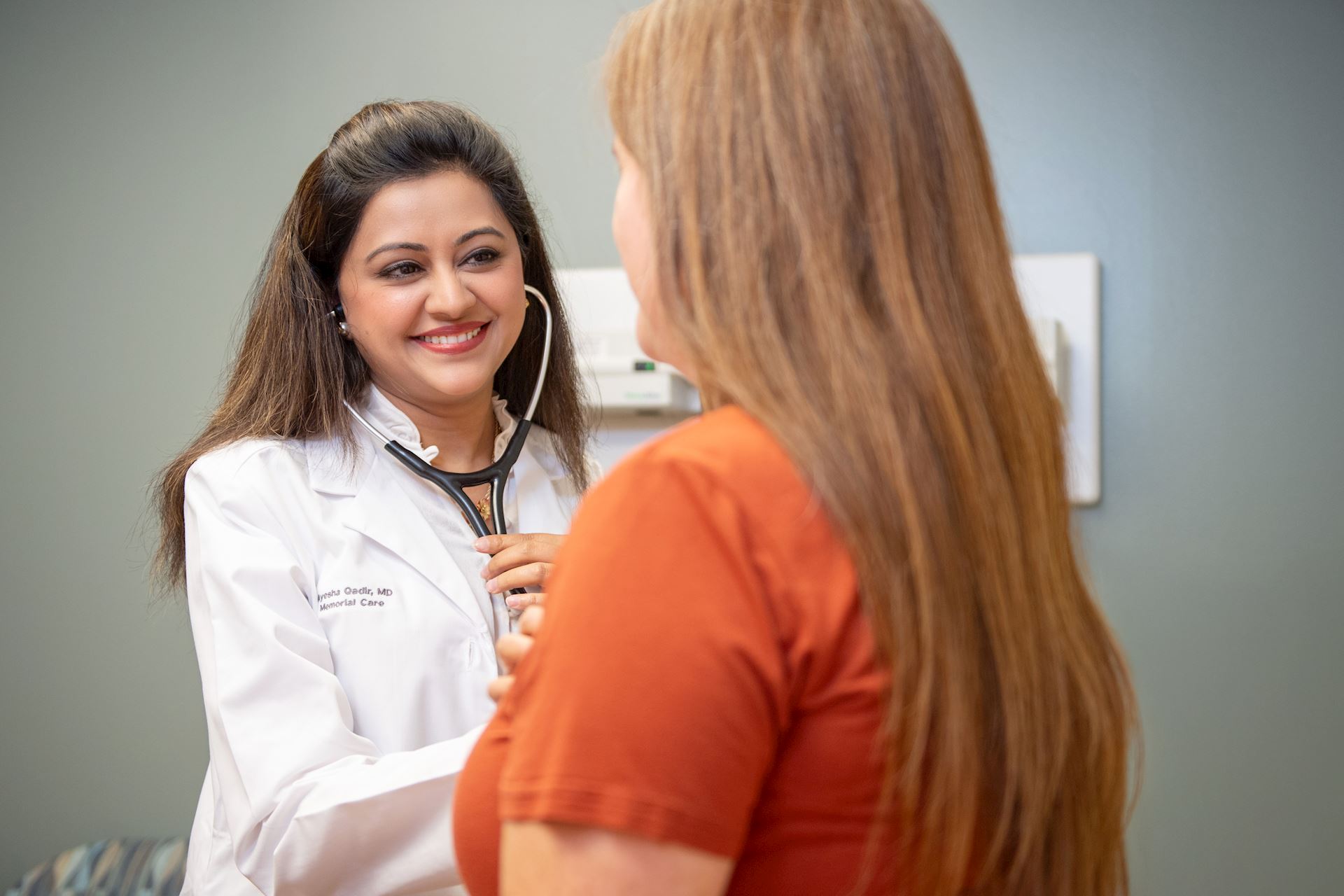 Photograph of a healthcare professional using a stethoscope to examine a patient in a clinical setting. The professional wears a white coat, and the patient is dressed in an orange shirt, highlighting a routine medical checkup scenario.