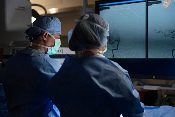 Photograph showing two medical professionals in surgical attire observing detailed vascular images on multiple monitors during a procedure. The scene highlights use of advanced imaging technology for real-time guidance in minimally invasive surgery.